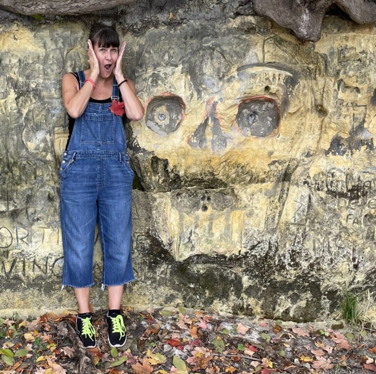 Photo of MB standing by an image of a skull carved into the rock, doing the "Home Alone" (oh no!) pose.